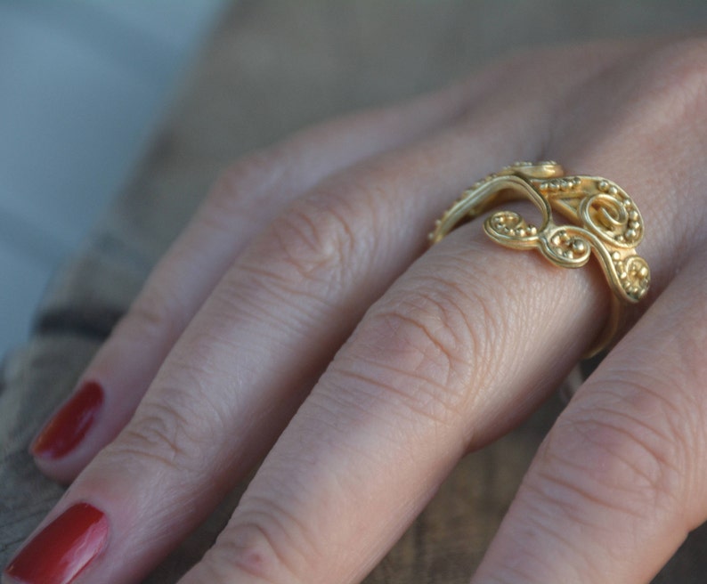 May include: A gold ring with a delicate, intricate design featuring swirling patterns and small beads. The ring is worn on a finger with red nail polish.