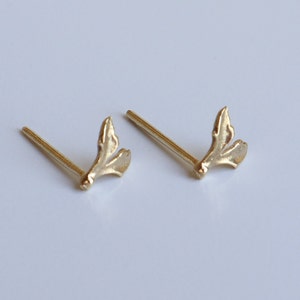 May include: A pair of gold-toned stud earrings featuring a small, organic, leaf-like or branch-inspired design. Each earring consists of a thin post attached to a textured, bifurcated metal shape that resembles a tiny sprout or botanical element.