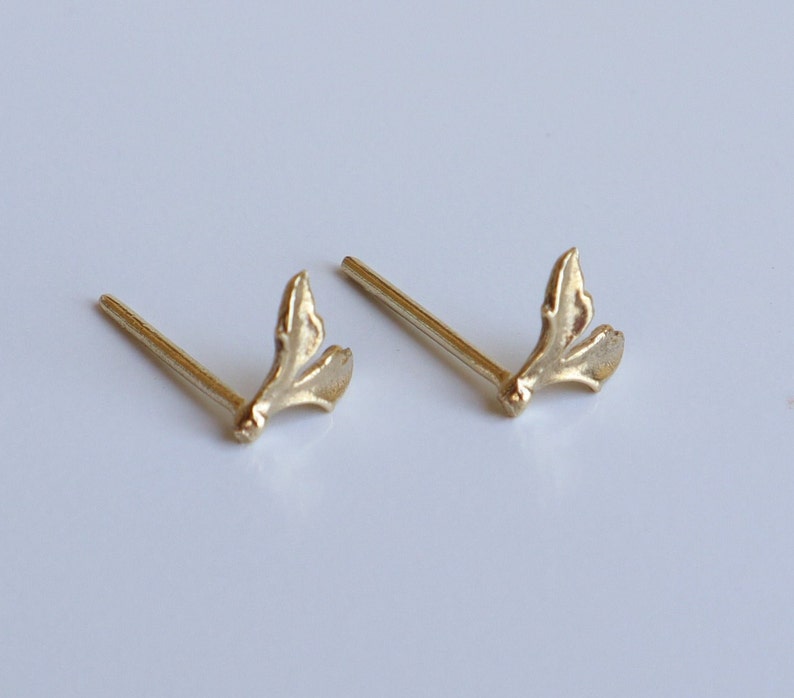 May include: A pair of gold-toned stud earrings featuring a small, organic, leaf-like or branch-inspired design. Each earring consists of a thin post attached to a textured, bifurcated metal shape that resembles a tiny sprout or botanical element.
