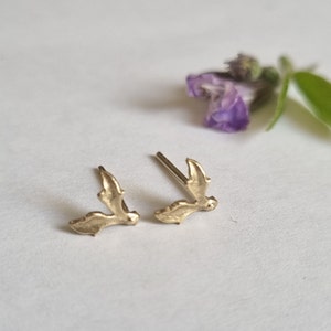 May include: A pair of gold-toned stud earrings shaped like small holly leaves. Each earring features a textured surface and a straight metal post extending from the back, designed for pierced ears. The earrings have a metallic finish.