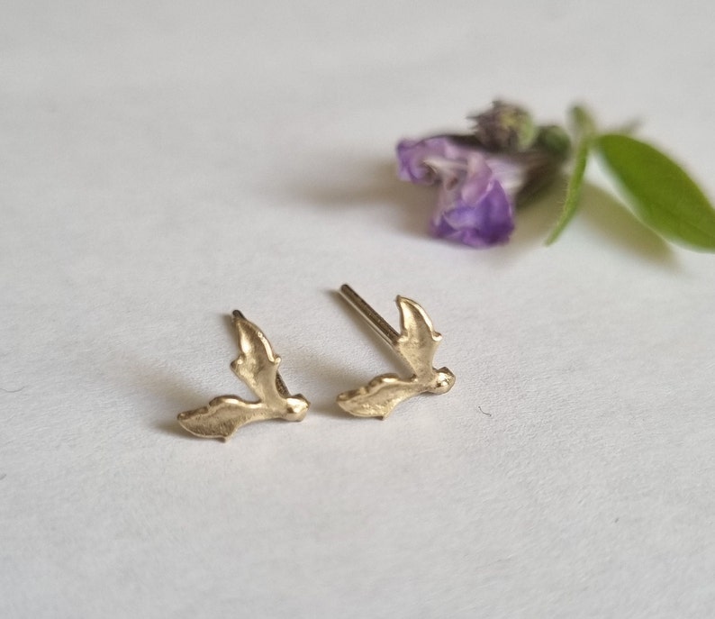 May include: A pair of gold-toned stud earrings shaped like small holly leaves. Each earring features a textured surface and a straight metal post extending from the back, designed for pierced ears. The earrings have a metallic finish.