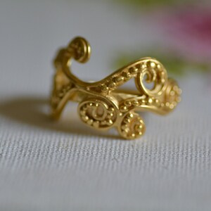 May include: A gold ring with a delicate, intricate design featuring swirling patterns and small beads.