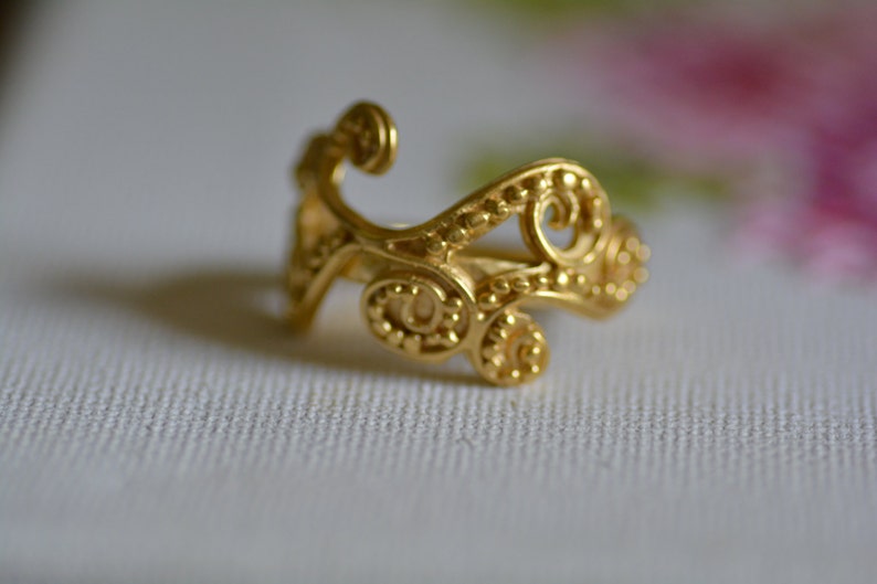 May include: A gold ring with a delicate, intricate design featuring swirling patterns and small beads.