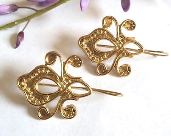 May include: A pair of gold-colored, ornate earrings in a mask shape. The earrings have a delicate, swirling design with small, raised details. A sprig of purple flowers is in the background.
