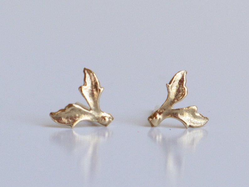May include: A pair of gold-toned stud earrings shaped like stylized holly leaves. Each earring features a textured, metallic surface with two distinct, pointed leaf segments extending from a central point, creating a botanical silhouette.
