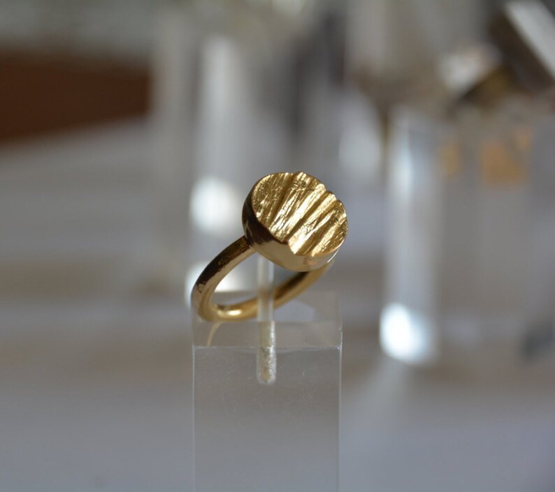May include: A gold ring with a textured, round, circular design. The ring is on a clear display stand.