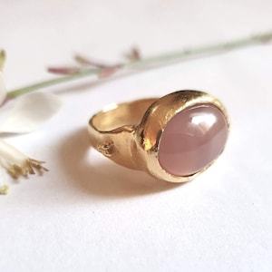 Puede incluir: Un anillo de oro con una gran piedra preciosa rosa de forma ovalada. El anillo tiene una banda texturizada.