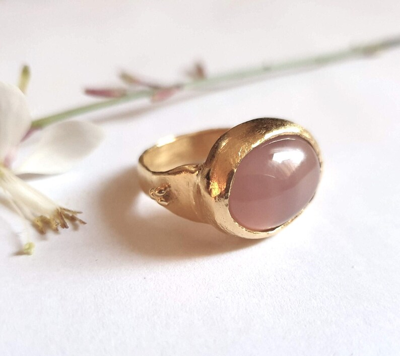 Puede incluir: Un anillo de oro con una gran piedra preciosa rosa de forma ovalada. El anillo tiene una banda texturizada.
