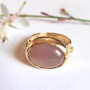 Puede incluir: Un anillo de oro con una piedra rosa ovalada grande engastada en un engaste de bisel. El anillo tiene una banda de oro texturizada.