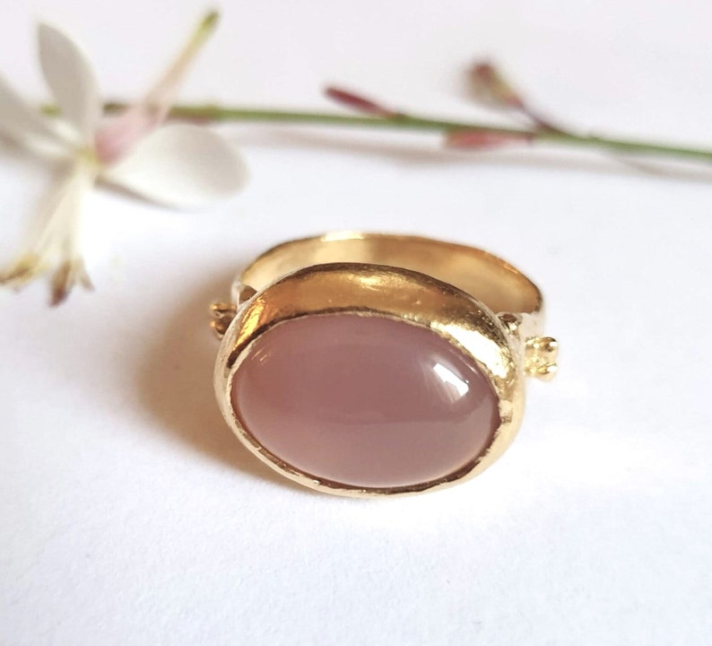 Puede incluir: Un anillo de oro con una piedra rosa ovalada grande engastada en un engaste de bisel. El anillo tiene una banda de oro texturizada.