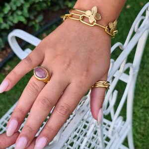 Puede incluir: Un brazalete de oro con una cadena delicada y detalles de flores. Un anillo con una piedra preciosa ovalada grande se lleva en el dedo &iacute;ndice.