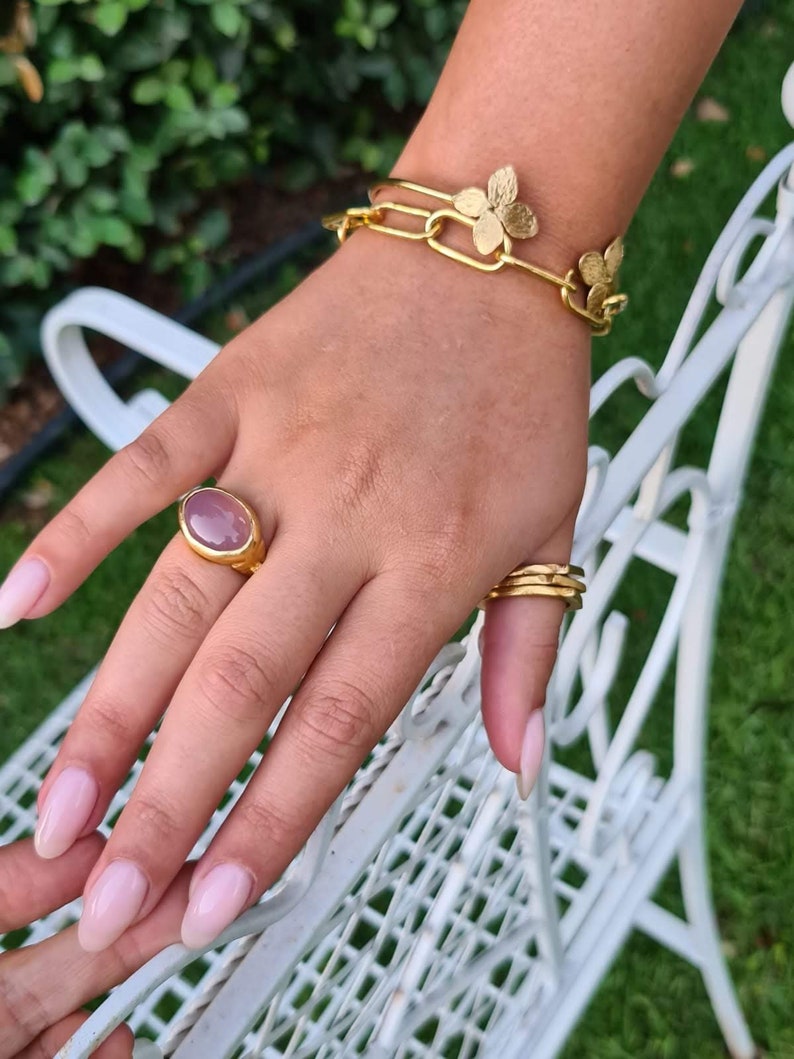 Puede incluir: Un brazalete de oro con una cadena delicada y detalles de flores. Un anillo con una piedra preciosa ovalada grande se lleva en el dedo &iacute;ndice.