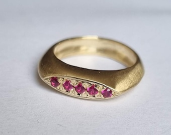 May include: A gold ring with a brushed finish, featuring six vibrant pink gemstones. The ring has a rounded band and an oval face. The gemstones are set in a row, adding a touch of colour to the gold.