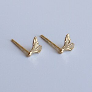 May include: A pair of gold-toned stud earrings featuring a small, textured leaf-like design. Each earring consists of a thin, straight post attached to a delicate, organic-shaped element that resembles a split leaf or a small botanical sprig.