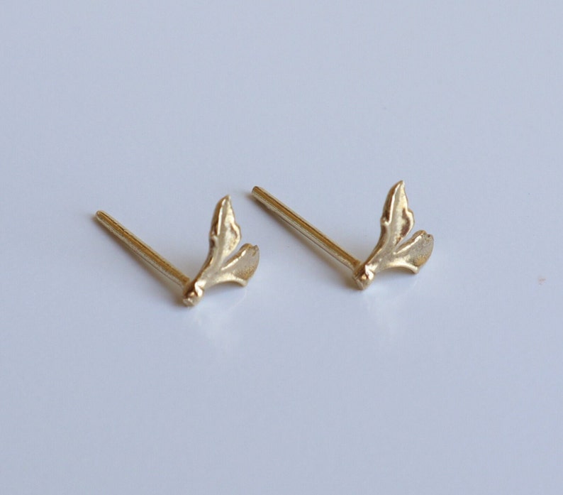 May include: A pair of gold-toned stud earrings featuring a small, textured leaf-like design. Each earring consists of a thin, straight post attached to a delicate, organic-shaped element that resembles a split leaf or a small botanical sprig.