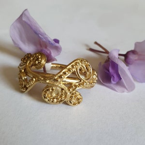 May include: A gold ring with a delicate, intricate design featuring swirling, interconnected loops and small beads. The ring is set against a white background with purple flower petals.