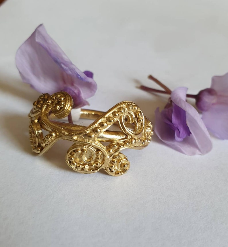 May include: A gold ring with a delicate, intricate design featuring swirling, interconnected loops and small beads. The ring is set against a white background with purple flower petals.