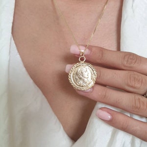May include: A gold chain necklace with a large, round pendant featuring a detailed profile of a person. The pendant has a rope-like border.
