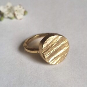May include: A gold ring with a textured, round face. The ring is made of a smooth metal and has a simple design.