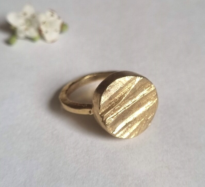 May include: A gold ring with a textured, round face. The ring is made of a smooth metal and has a simple design.