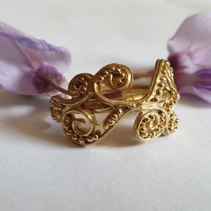 May include: A gold ring with a delicate, intricate design featuring swirling patterns and small beads. The ring is made of metal and has a polished finish.