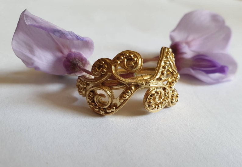 May include: A gold ring with a delicate, intricate design featuring swirling patterns and small beads. The ring is made of metal and has a polished finish.