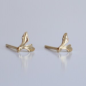 May include: A pair of gold-toned stud earrings featuring a delicate, organic branch-like design with two small, leaf-shaped protrusions extending from a central stem. Each earring includes a straight post extending from the back for secure attachment.