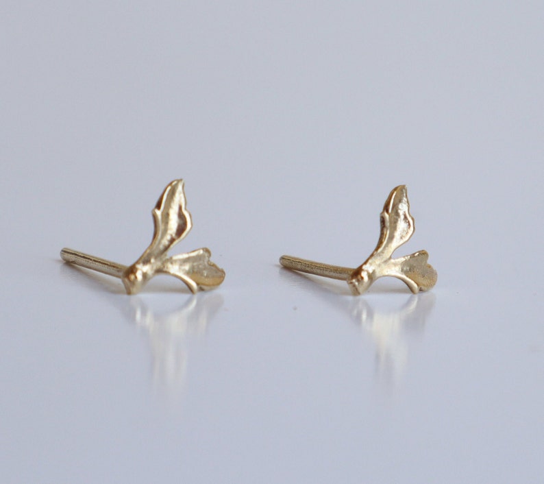 May include: A pair of gold-toned stud earrings featuring a delicate, organic branch-like design with two small, leaf-shaped protrusions extending from a central stem. Each earring includes a straight post extending from the back for secure attachment.