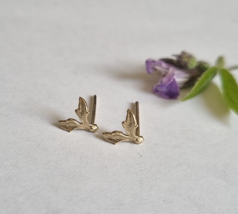 May include: A pair of gold-toned stud earrings shaped like small, textured leaves. Each earring features a delicate, organic silhouette with a post backing, designed to sit flush against the earlobe with a subtle, metallic sheen and detailed edges.