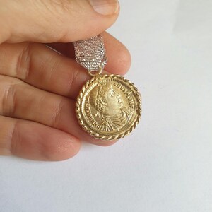 May include: A gold-colored pendant with a rope-like border and a profile image of a person with a crown. The pendant is hanging from a silver and white ribbon.