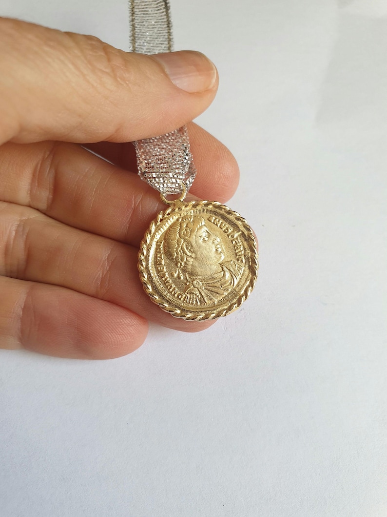 May include: A gold-colored pendant with a rope-like border and a profile image of a person with a crown. The pendant is hanging from a silver and white ribbon.