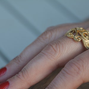 May include: A gold ring with a delicate, intricate design. The ring is worn on a finger with red nail polish.