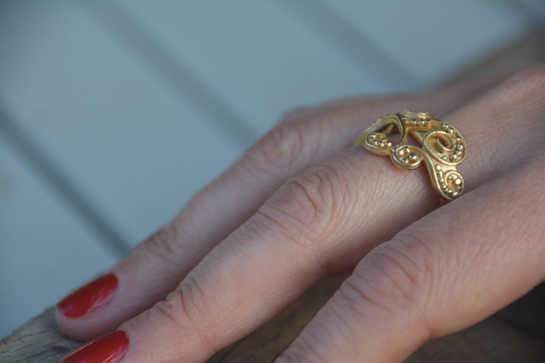 May include: A gold ring with a delicate, intricate design. The ring is worn on a finger with red nail polish.