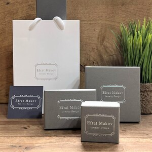 May include: Four gray boxes with the text "Efrat Makov Jewelry Design" on them. The boxes are stacked on top of each other, with the largest box on the bottom and the smallest box on top. The boxes are sitting on a wooden surface.