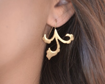 May include: A close-up of a gold-colored earring with a floral design. The earring is worn on a person's ear. The earring has a hook and a decorative, stylized floral shape with curved lines and rounded edges. The background is blurred.