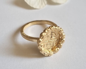 May include: A gold ring with a textured, circular design featuring a raised, detailed image. The ring is made of metal and has a simple band.