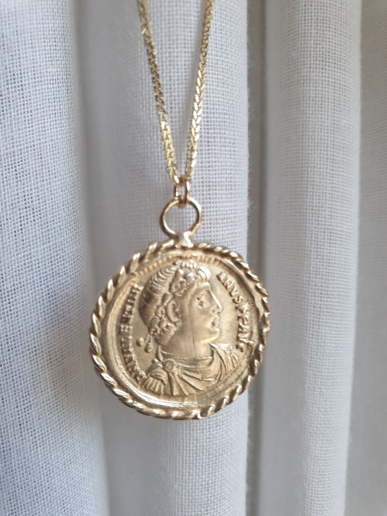 May include: A gold chain necklace with a round pendant featuring a profile of a person with a crown. The pendant has a rope-like border and the inscription "IMP. CAES. FL. ANN. VERUS. AUG. P. F. A." around the edge.