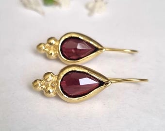 May include: A pair of gold-colored earrings with a teardrop-shaped, deep red gemstone. The earrings have a decorative cluster of small gold beads at the top and a simple hook closure. The background is a soft, neutral color.