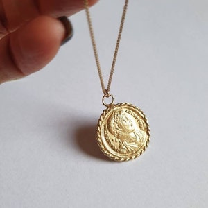 May include: A gold chain necklace with a round pendant featuring a profile of a woman's face. The pendant has a rope-like border.