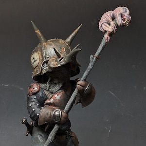 Peut inclure: Une petite figurine détaillée d'un guerrier portant un casque avec des cornes et tenant un bâton surmonté d'une créature. La figurine est peinte dans des tons de gris, de brun et de rouge.