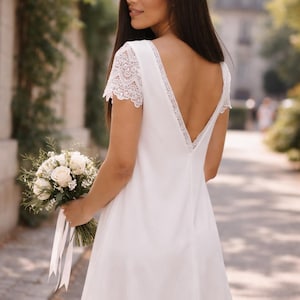 May include: A white, short-sleeved dress with lace detailing on the sleeves and neckline. The dress has a V-neck back and is paired with a bouquet of white roses and greenery. The dress is a simple, elegant design.