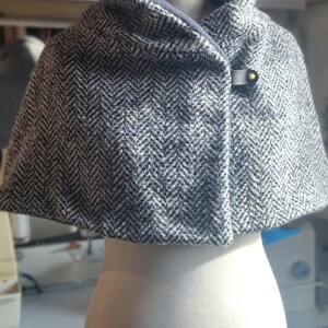 May include: A gray and white herringbone pattern cape with a navy blue collar and lining. The cape is fastened with a leather strap and a gold-colored button. The garment is displayed on a white mannequin.