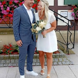 May include: A couple smiles, posing outdoors. The man wears a blue suit and white sneakers. The woman wears a white dress and silver heels, holding a bouquet. Pink heart balloons float above them. The text "ROBE MARI&Eacute;E COURTE" is at the bottom.
