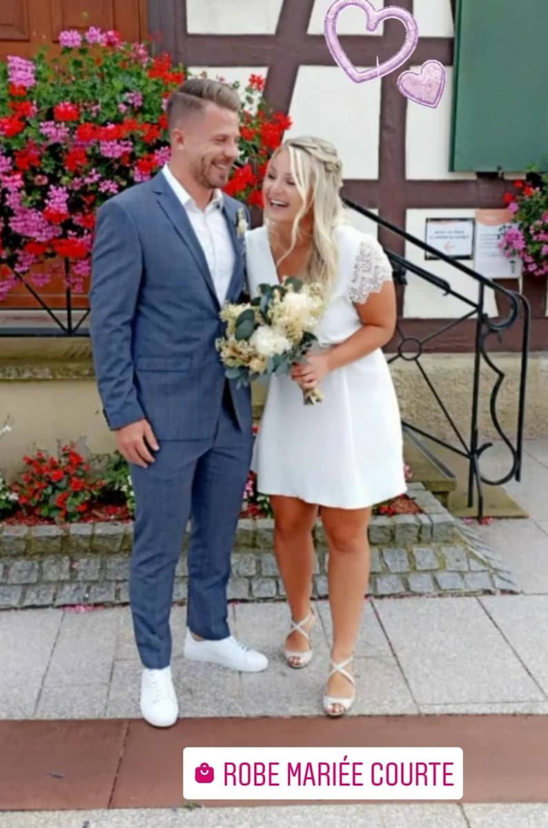 May include: A couple smiles, posing outdoors. The man wears a blue suit and white sneakers. The woman wears a white dress and silver heels, holding a bouquet. Pink heart balloons float above them. The text "ROBE MARI&Eacute;E COURTE" is at the bottom.