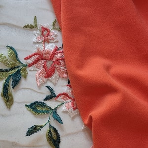 Pu&ograve; includere: Primo piano di un tessuto bianco con ricami floreali in tonalit&agrave; di verde e rosa. Un pezzo di tessuto arancione &egrave; drappeggiato sul tessuto bianco.
