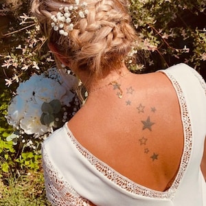 May include: A woman's hairstyle features a braided updo adorned with small white flowers. She wears a white dress with lace detailing and a gold necklace. A bouquet of white flowers is held in front of her.