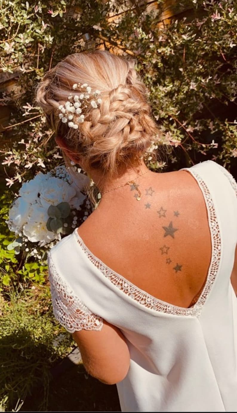 May include: A woman's hairstyle features a braided updo adorned with small white flowers. She wears a white dress with lace detailing and a gold necklace. A bouquet of white flowers is held in front of her.