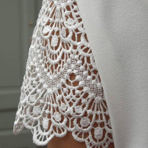 May include: Close-up of a white dress sleeve featuring intricate lace detailing. The lace has a scalloped edge and a delicate, openwork design. The sleeve is paired with a smooth, solid white fabric.