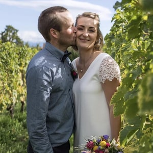 May include: A couple in a vineyard, the woman in a white dress with lace sleeves, holding a bouquet of colorful flowers. The man wears a blue button-down shirt and a bow tie, kissing her cheek. The background shows green grapevines.