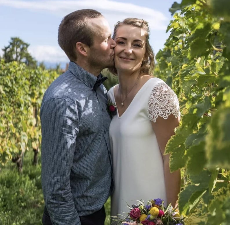 May include: A couple in a vineyard, the woman in a white dress with lace sleeves, holding a bouquet of colorful flowers. The man wears a blue button-down shirt and a bow tie, kissing her cheek. The background shows green grapevines.